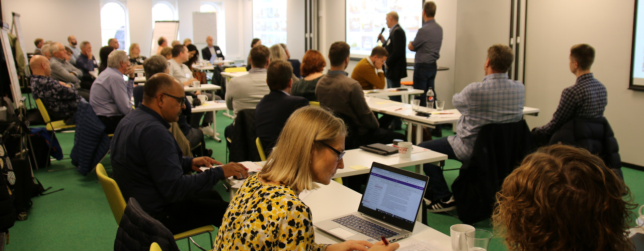 Samling mennesker på seminar sitter ved bord og skriver