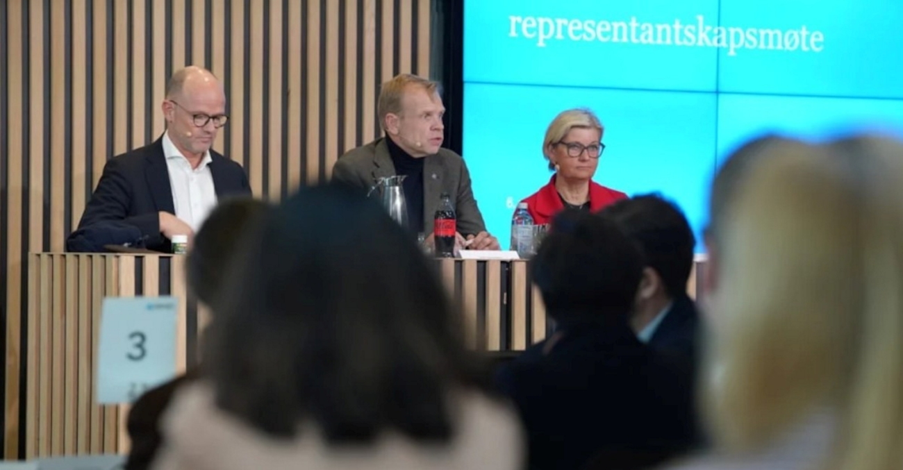 Tre mennesker sittende på en scene med publikum foran.