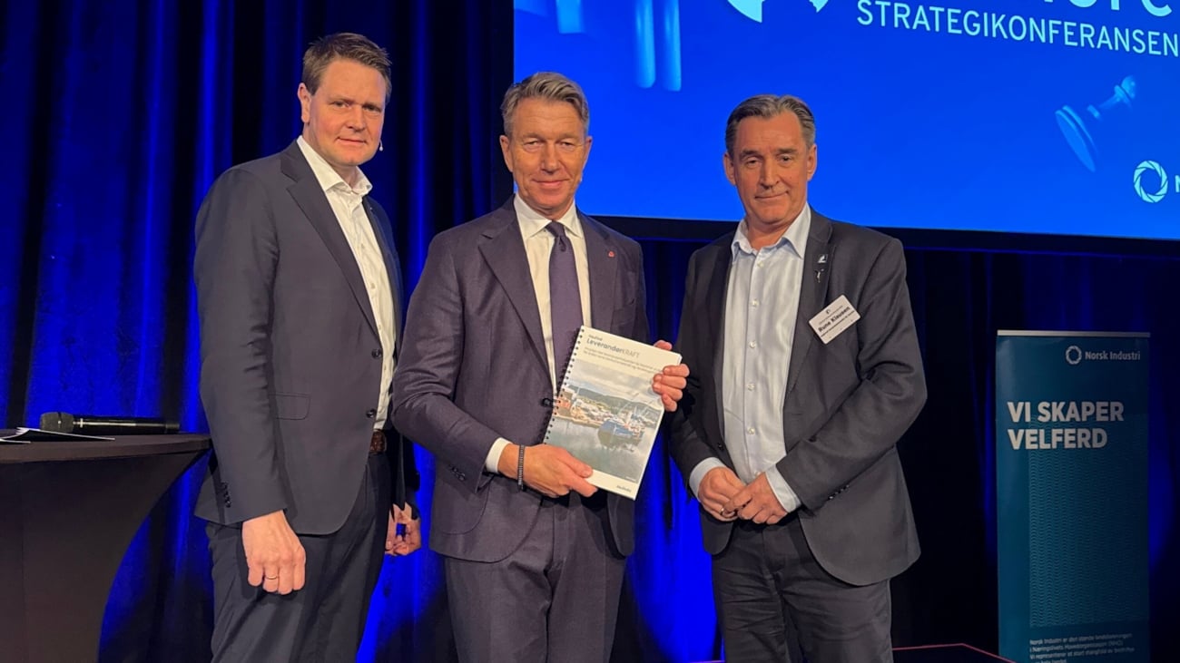 Tre menn på en scene i forbindelse med overlevering av en rapport.